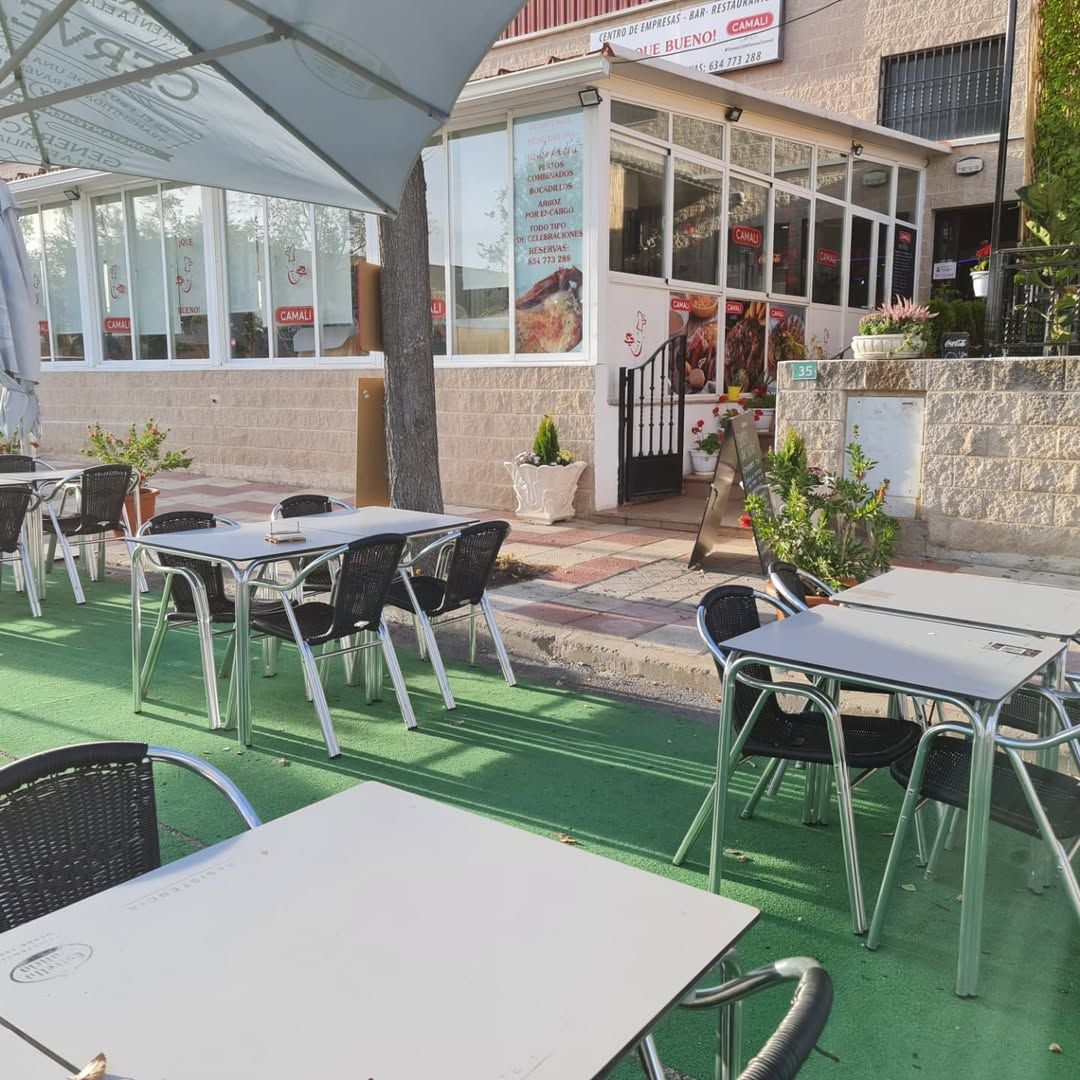 Zona de restaurante al aire libre bajo techo de metal, con mesas, sillas y plantas decorativas.