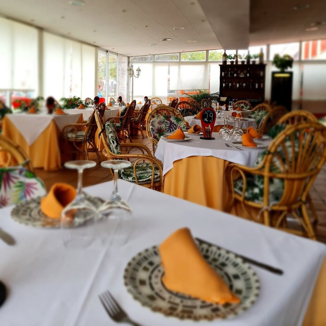 Interior de restaurante con mesas preparadas para comer, sillas de mimbre, manteles blancos y servilletas naranjas.