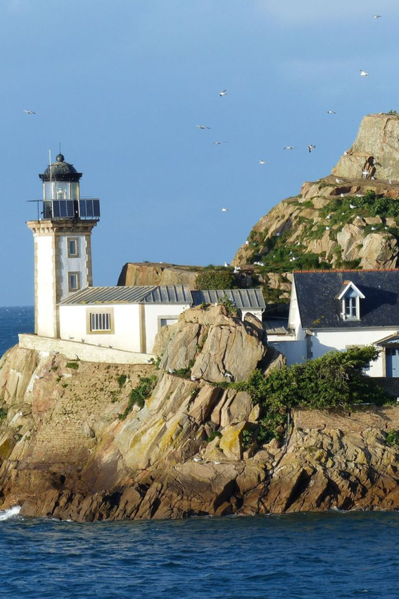 Un phare et des bâtiments blancs se trouvent sur une île rocheuse, l'océan en face, le ciel bleu au-dessus.