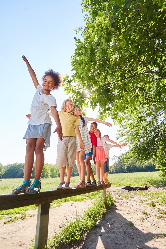 Enfants en équilibre sur une poutre en bois à l'extérieur, l'un avec le bras levé, journée ensoleillée.