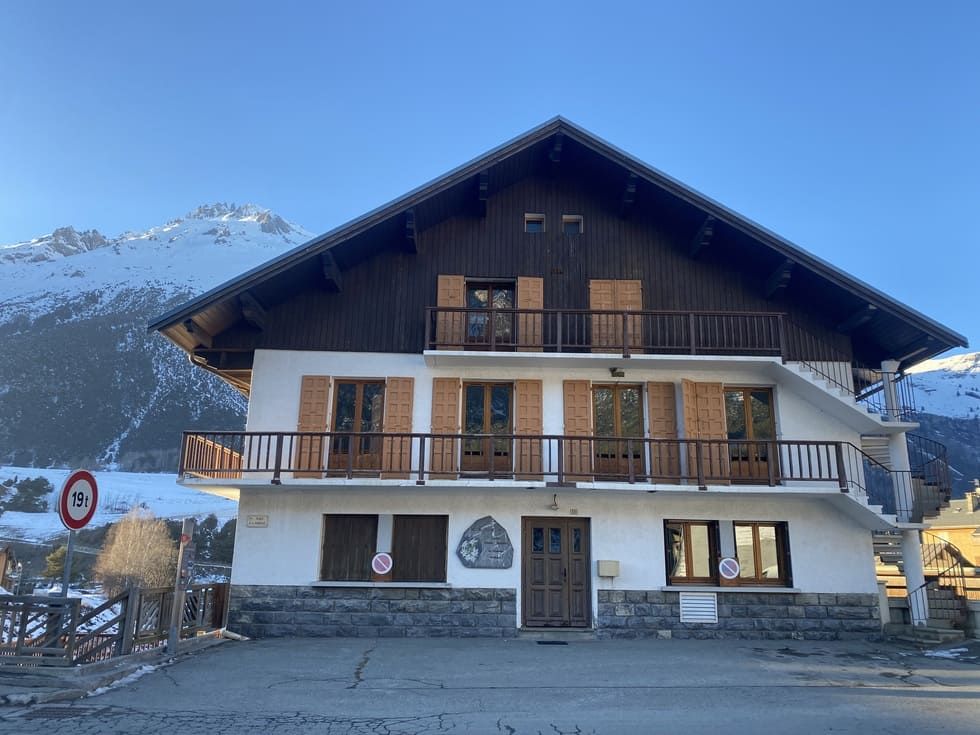 Bâtiment en bois de deux étages avec volets lumineux. Montagnes enneigées en arrière-plan.