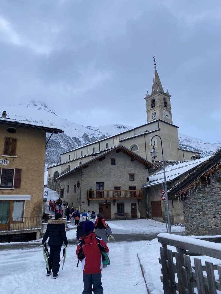 Scène de rue enneigée devant une église avec un haut clocher, des gens marchant avec des vêtements de neige et des sacs à dos.