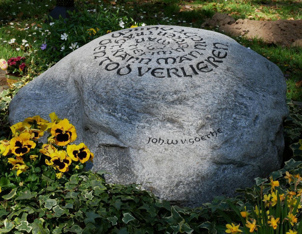 Großer, gravierter grauer Stein in einem Blumengarten mit gelben Blüten und Grünpflanzen.