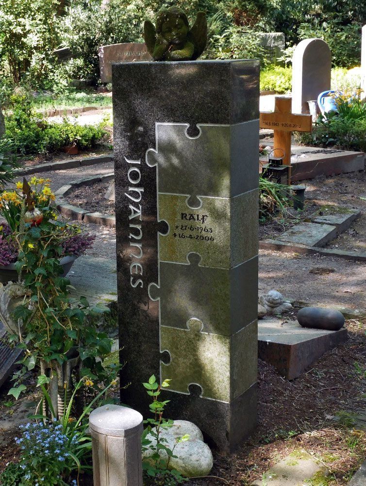 Eine Gedenksäule in Form eines Puzzles in einem Garten, dahinter Blumen, Steine ​​und ein Kreuz.