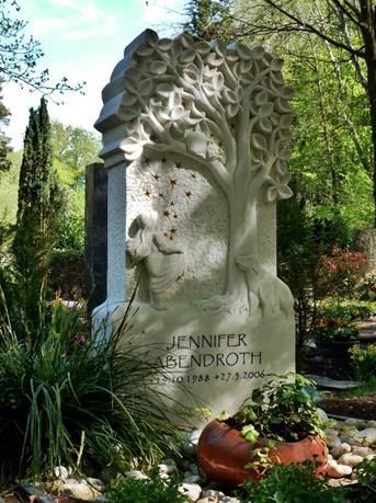 Weißer, baumförmiger Grabstein mit floralen Verzierungen in einem üppigen Garten, beschriftet mit „Jennifer und John“, mit Kürbissen am Sockel