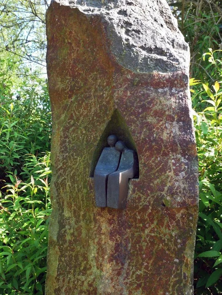 Kleine Bronzefigur, eingelassen in einer Aussparung eines hohen, moosbewachsenen Steins in einem grünen Garten.
