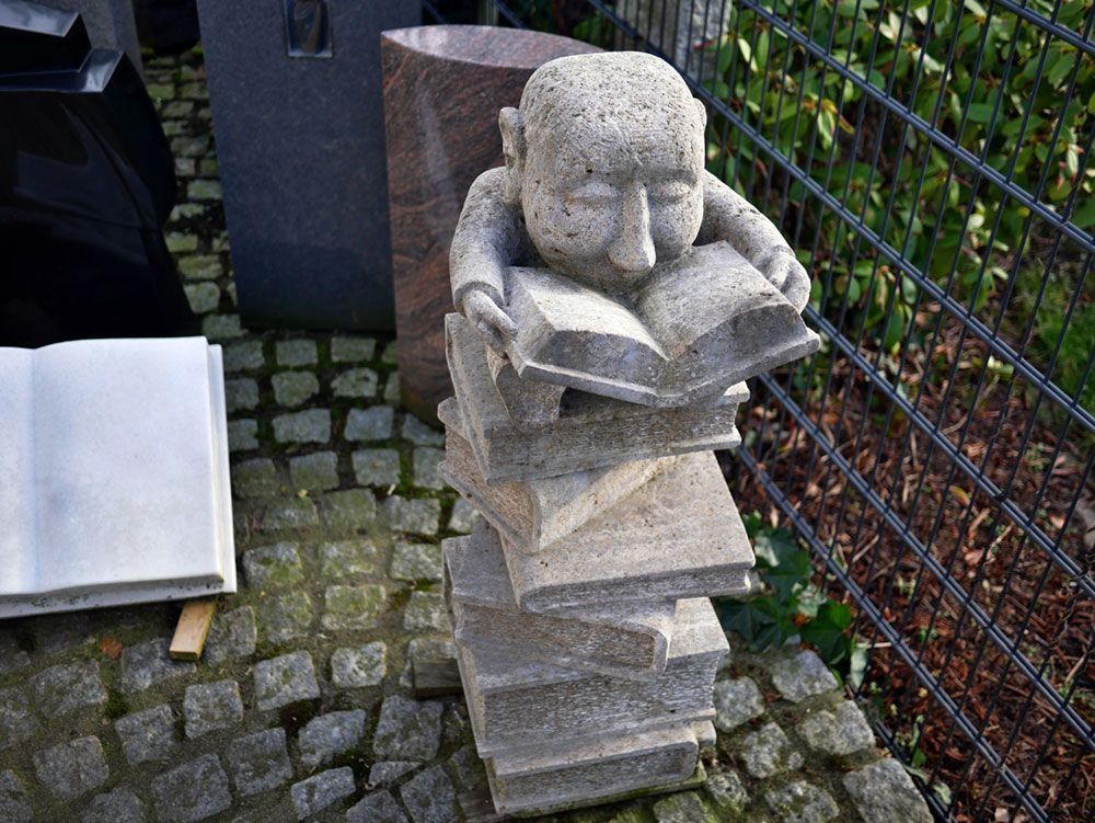 Steinskulptur eines Gesichts auf gestapelten Büchern neben einem Kopfsteinpflasterweg und einem Zaun