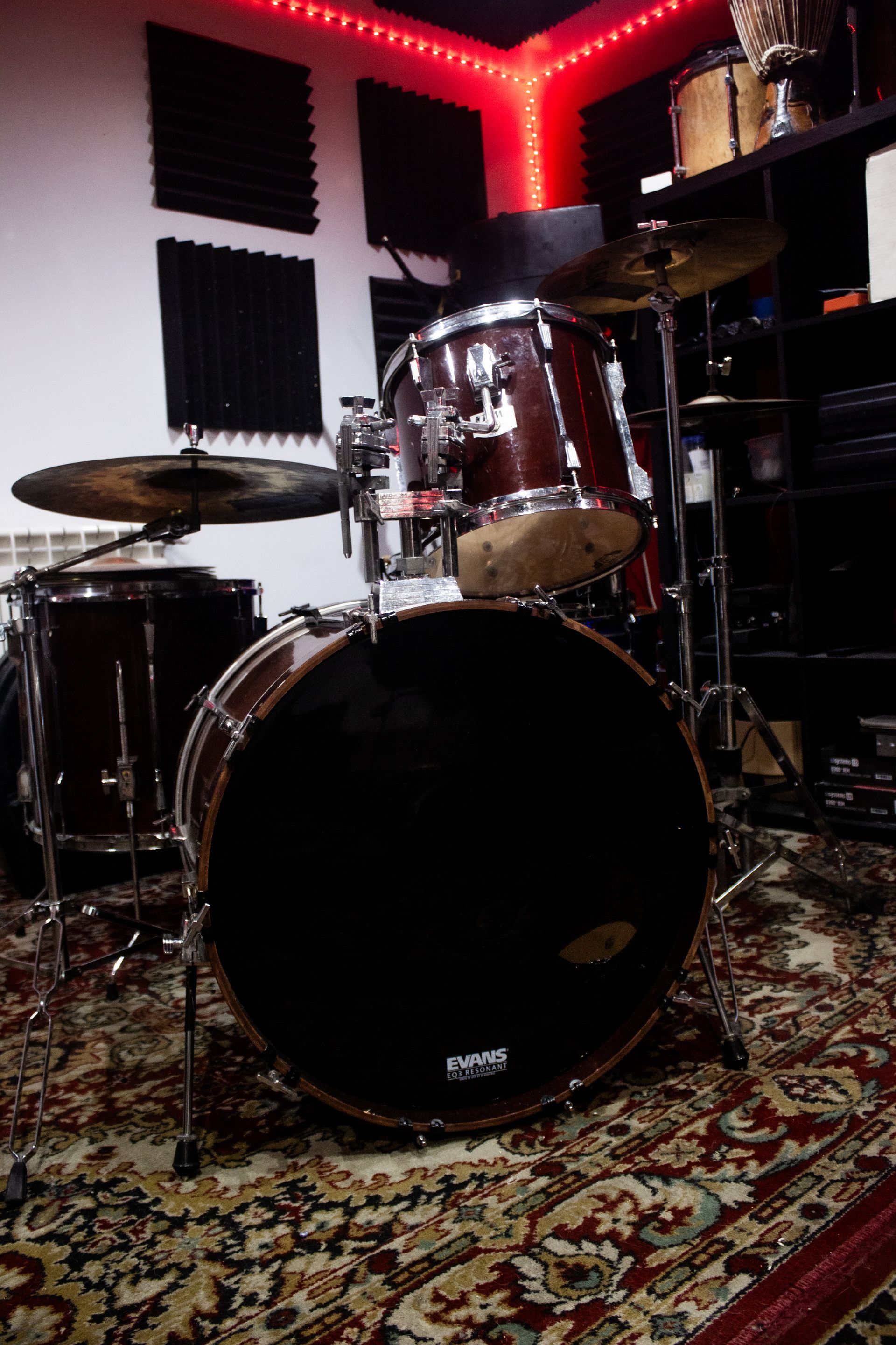 Batería en un estudio de música con iluminación roja cenital, rodeada de material insonorizante y equipos.
