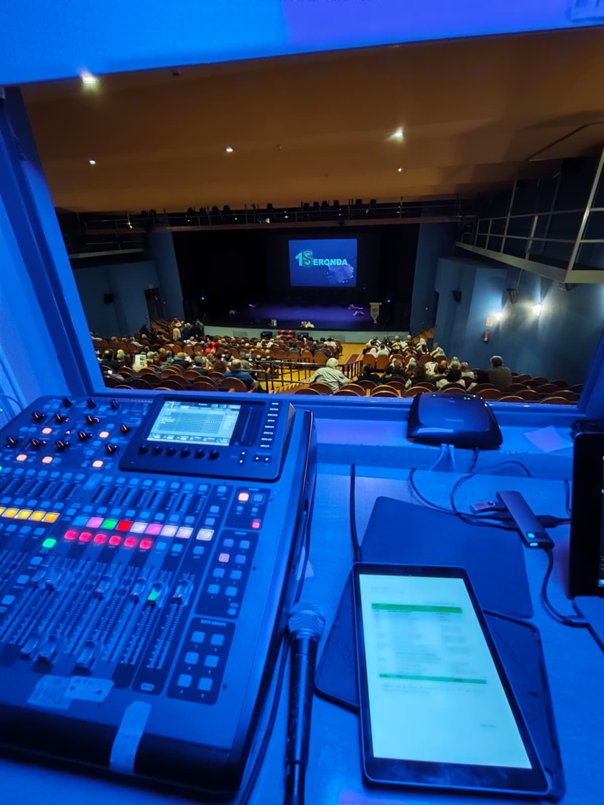 Vista desde una cabina de sonido: mesa de mezclas, teléfono inteligente y un teatro con público iluminado en azul.