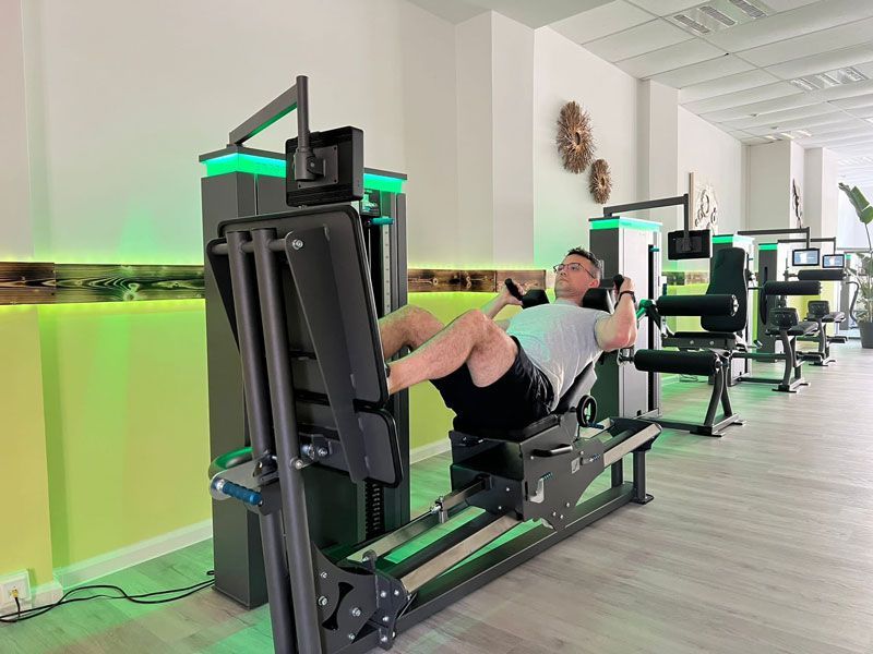 Ein Mann benutzt eine Beinpresse in einem Fitnessstudio. Weitere Geräte sind in einem hellen Raum zu sehen.