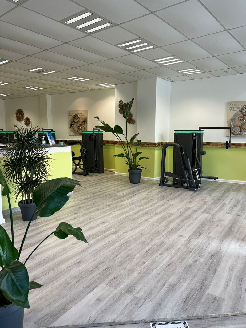 Das Fitnessstudio ist innen mit hellem Holzboden, Trainingsgeräten, Pflanzen und weißen Wänden mit grünen Akzenten ausgestattet.