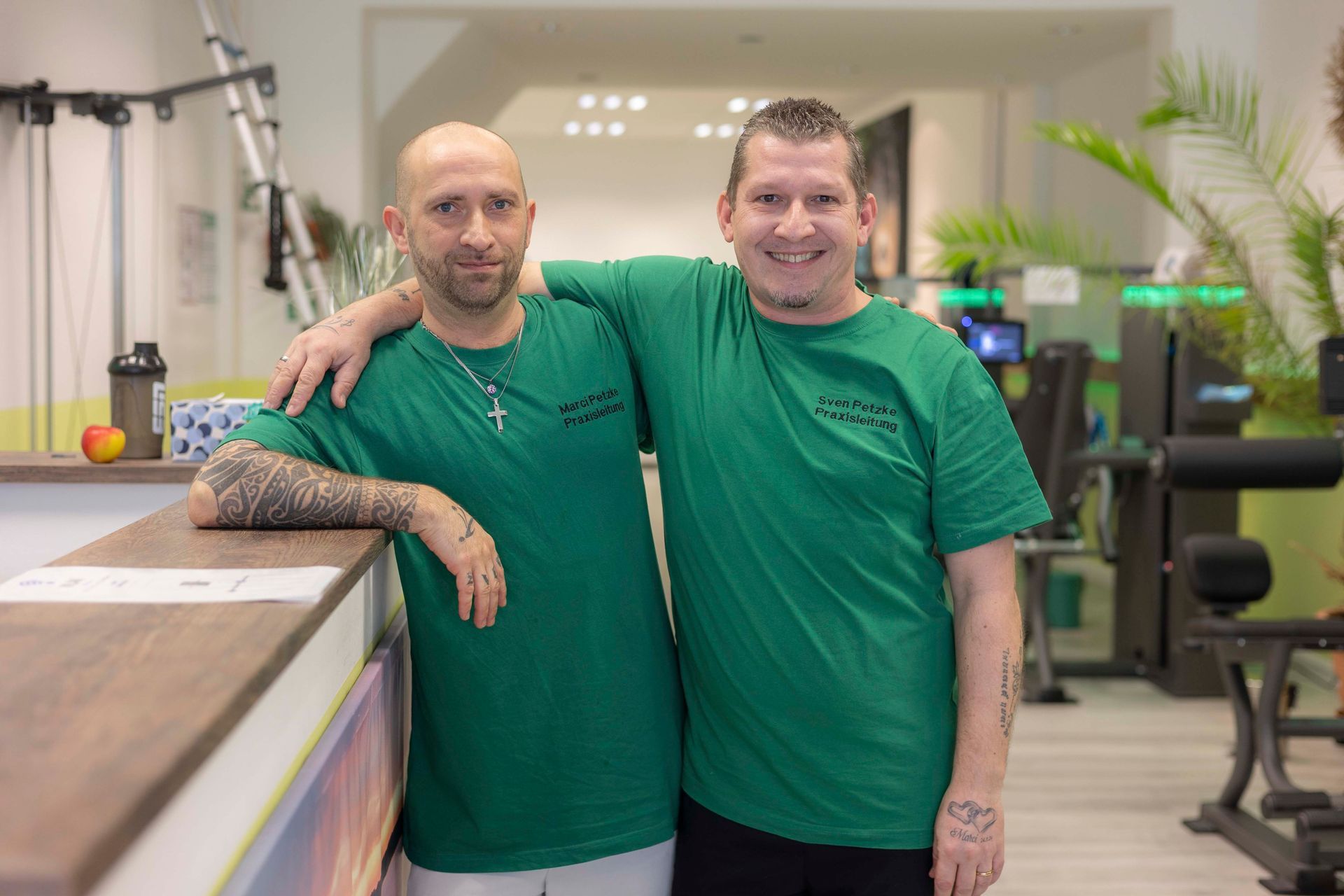 Zwei Männer in weißen Hemden umarmen sich lächelnd. Einer trägt eine Baseballkappe. Grüner Hintergrund.