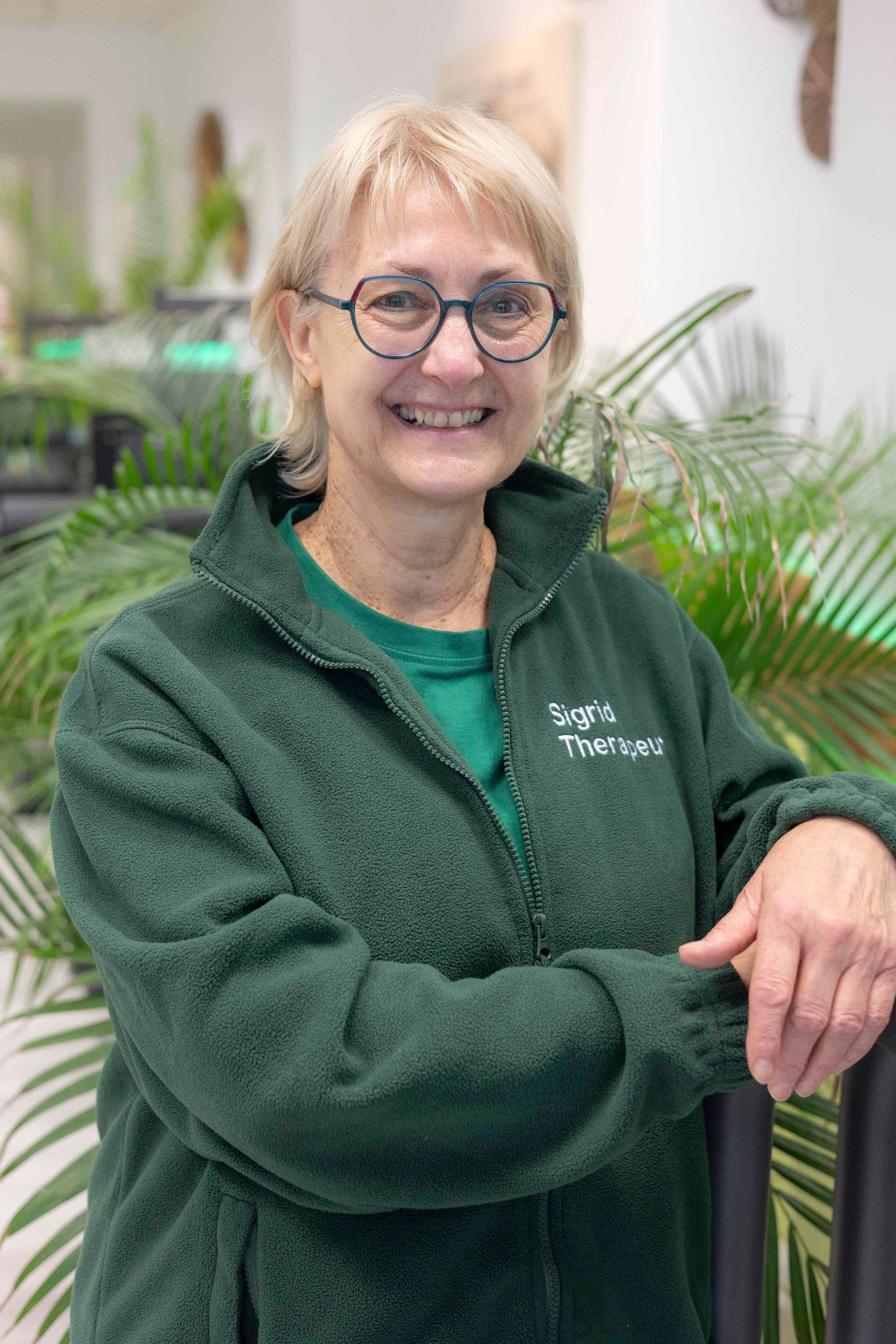 Frau mit kurzen blonden Haaren und Brille lächelt; sie trägt ein blaues Hemd vor grünem Hintergrund.