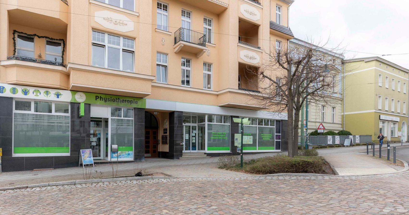Schaufenster einer Physiotherapiepraxis mit grünen Akzenten. Beiges Gebäude mit großen Fenstern und einem abfallenden Gehweg.
