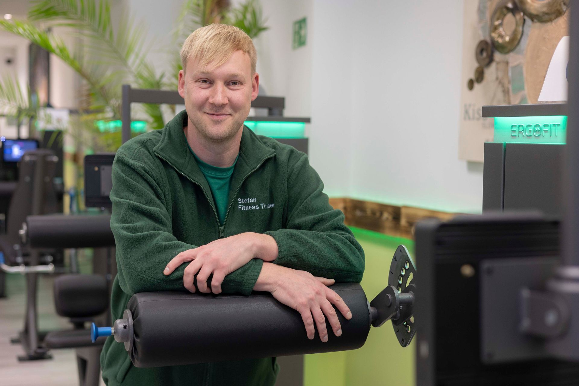 Eine blonde Person lächelt in die Kamera; sie trägt ein blaues T-Shirt mit V-Ausschnitt; der Hintergrund ist einfarbig grün.