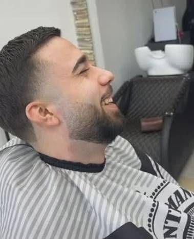 Ein Mann mit Bart sitzt in einem Friseurstuhl und lächelt. Foto vom Ado Barbershop, Inh. Bijo