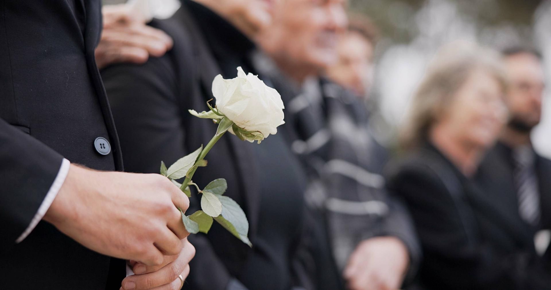 Ein Mann im Anzug hält bei einer Beerdigung eine weiße Rose in der Hand.