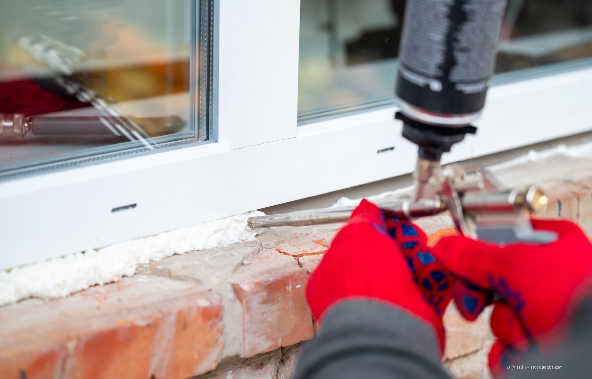 Person mit roten Handschuhen, die Sprühschaum verwendet, um einen Fensterrahmen an einer Ziegelwand abzudichten.