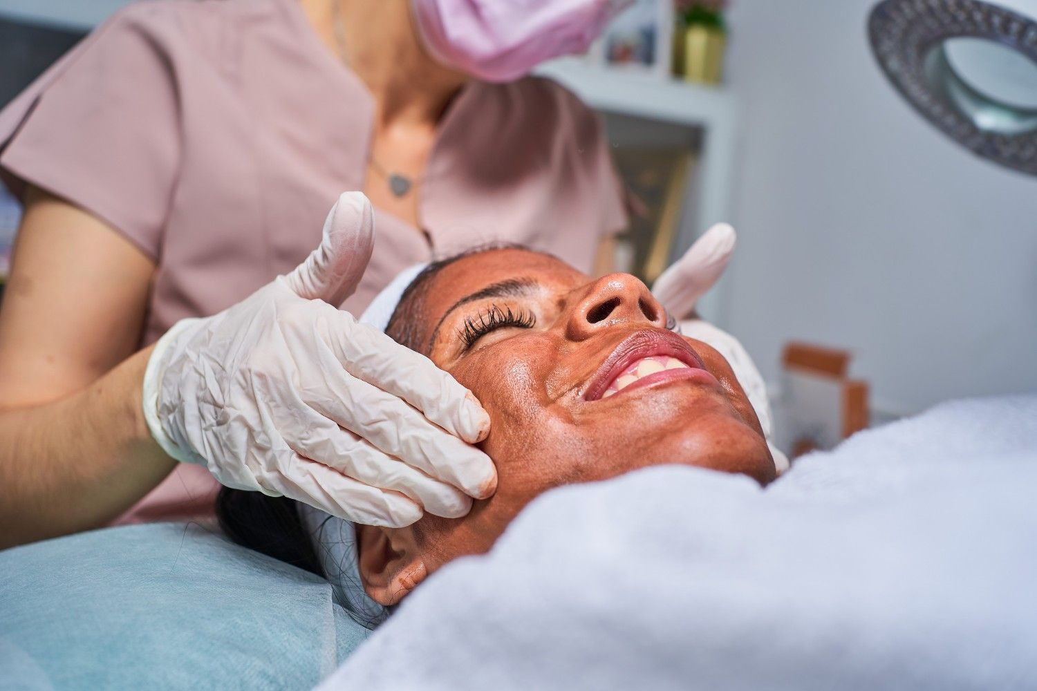 Mujer recibiendo tratamiento facial en un spa, terapeuta con guantes y mascarilla.