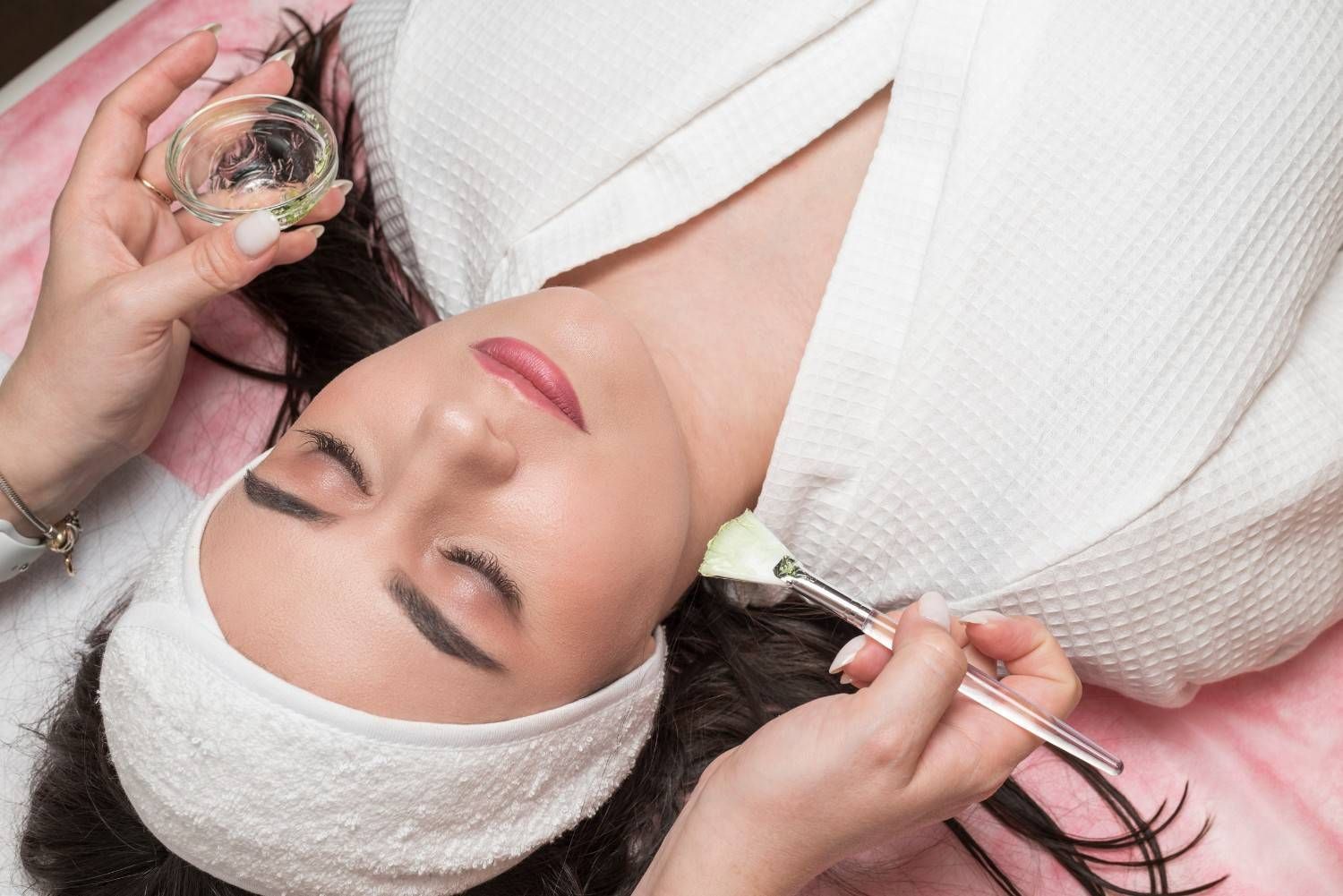 Mujer recibiendo tratamiento facial en un spa, tumbada con los ojos cerrados. Una esteticista le aplica una mascarilla.