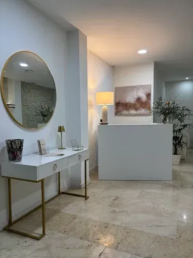 Recepción blanca y luminosa con escritorio, espejo y obras de arte. Detalles dorados, suelos de mármol y plantas.