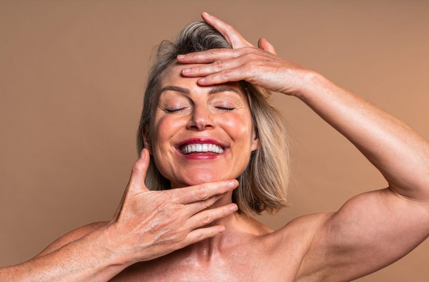 Una mujer de pelo corto y gris sonríe, tocándose la cara con ambas manos, con los ojos cerrados, sobre un fondo bronceado.