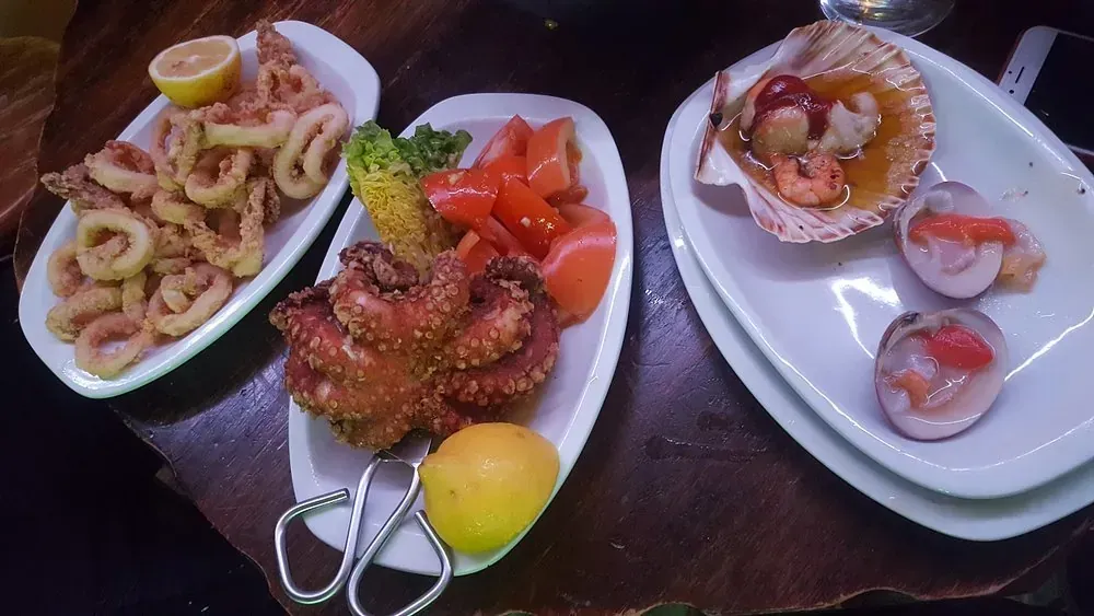 Platos de mariscos: calamares fritos, pulpo, vieiras y otros mariscos en platos blancos, sobre una mesa de madera.