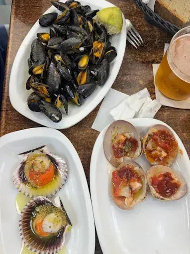 Platos de mariscos en platos blancos: mejillones, vieiras y almejas, con cerveza y rodaja de limón sobre una mesa de madera.