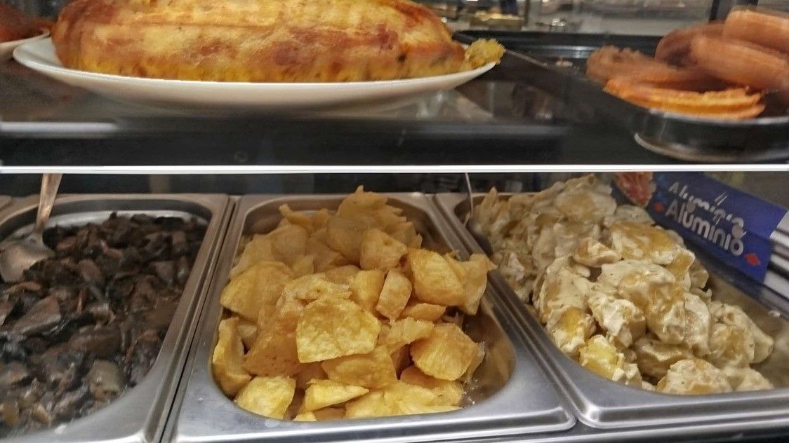 Bufé de autoservicio con bandejas de carnes asadas y patatas bajo una vitrina de cristal.