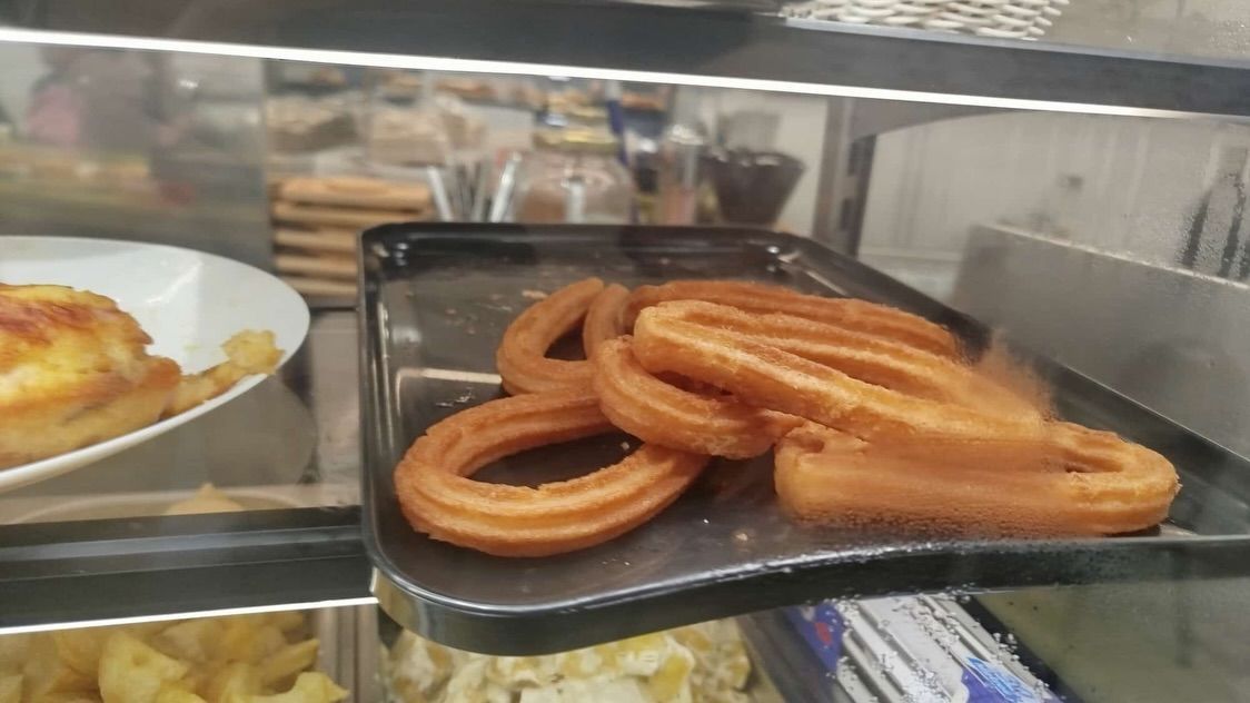 Bandeja de churros en una vitrina de panadería, con un pastel en un plato a la izquierda.