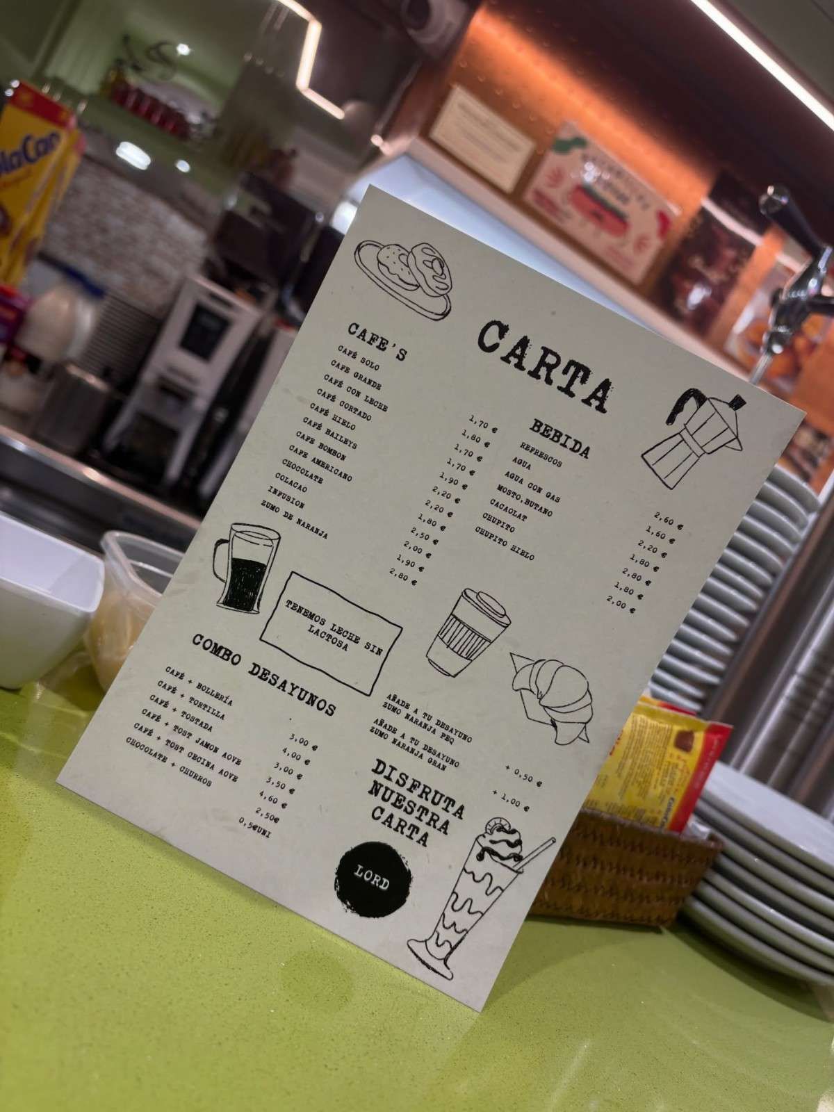 La carta del menú está sobre el mostrador de una cafetería, inclinada junto a un tarro de cristal y un servilletero a rayas.