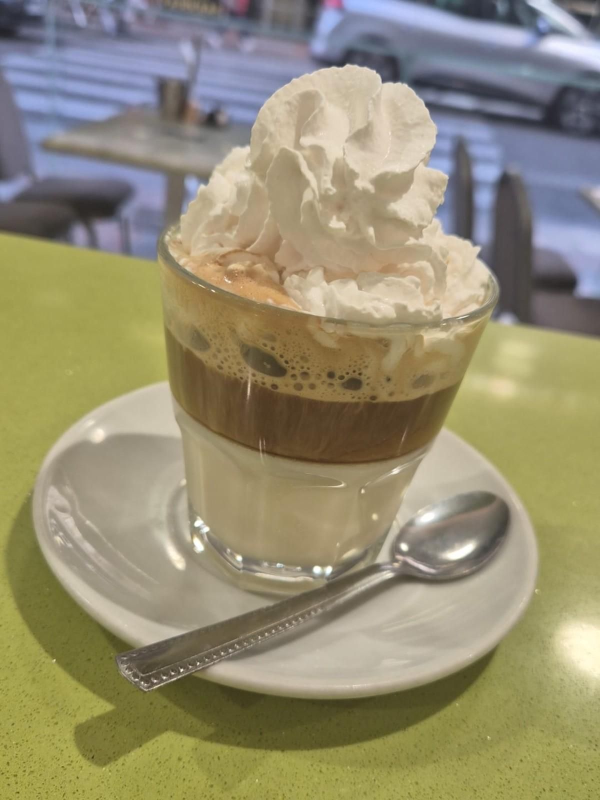 Café helado en capas con crema batida sobre un platillo, con una cuchara sobre la mesa de un café.