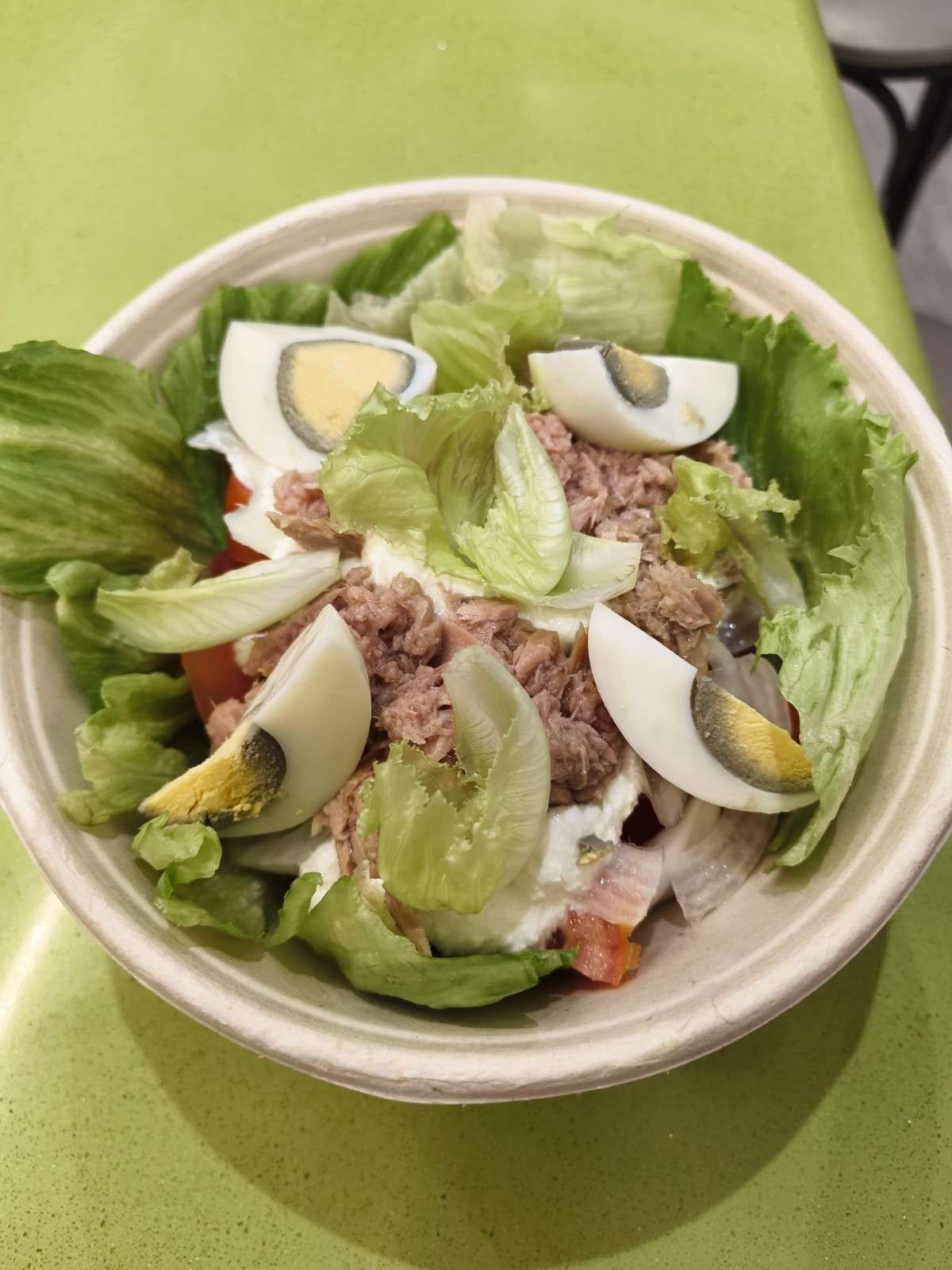 Bol de ensalada de atún con lechuga, rodajas de huevo, tomates y aderezo sobre una mesa verde.