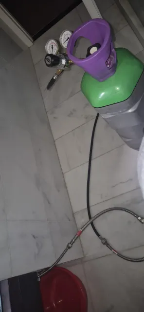 Cilindro de gas morado y verde con manguera y manómetro sobre suelo de baldosas.