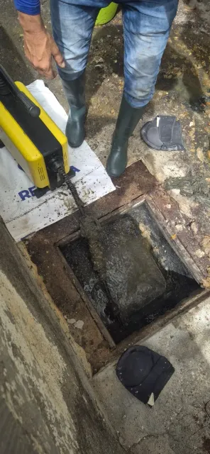 Pozo de registro de alcantarillado abierto con botas de trabajador y una herramienta de medición amarilla sobre un suelo de hormigón sucio.