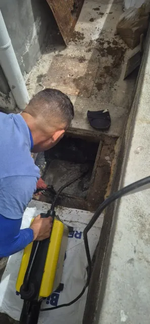 Técnico inspeccionando un desagüe de piso con una máquina desatascadora amarilla en un espacio de servicio estrecho.
