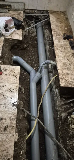 Zanja abierta con tuberías de PVC grises y escombros en una zona de construcción, que muestra trabajos de fontanería subterránea.