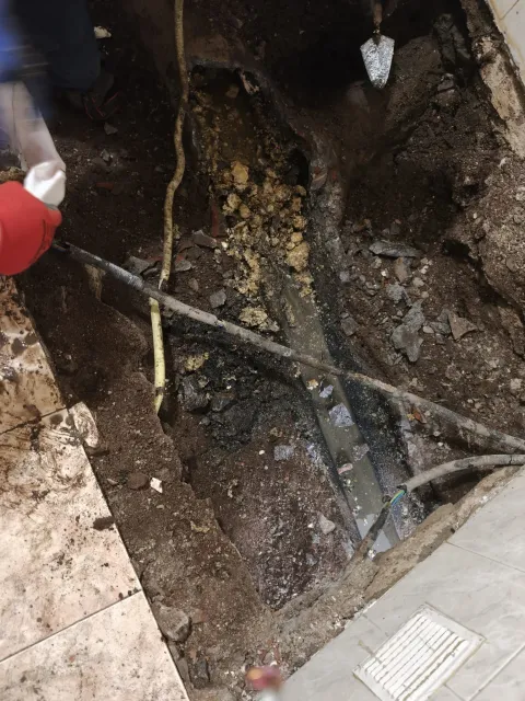 Persona que utiliza una herramienta para inspeccionar una zanja fangosa con tuberías y cables expuestos bajo tierra.
