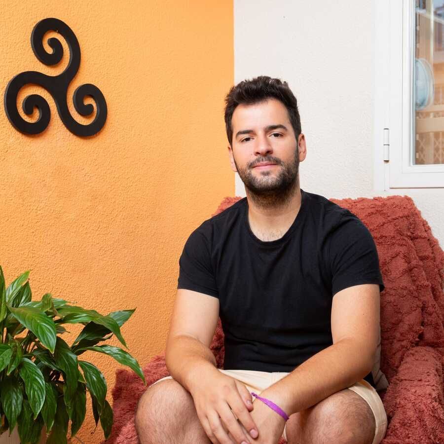 Un hombre con barba está sentado en un sillón rojo, con camisa negra y pantalones cortos color canela. Hay una planta y un símbolo en la pared.