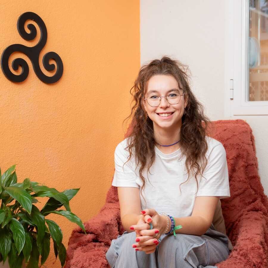 Una joven con gafas sonríe, sentada en una silla. Está en un interior, con una pared naranja y un símbolo decorativo de espiral.