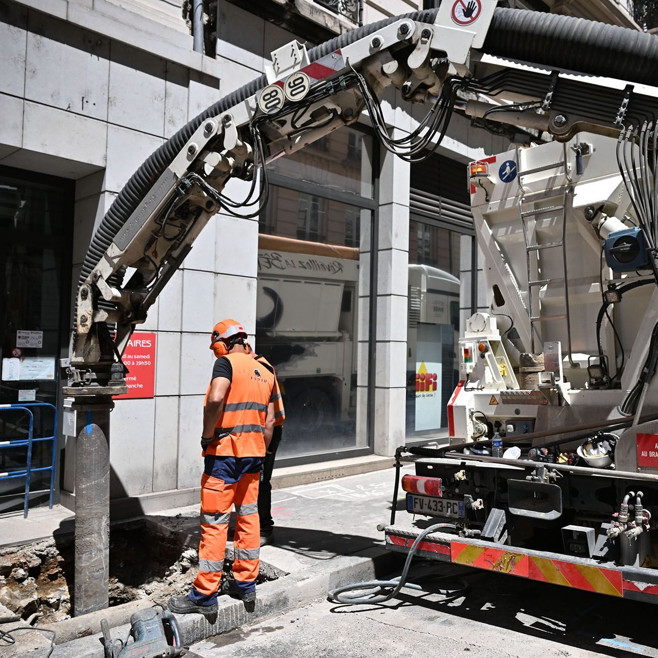 Un ouvrier vêtu d'une combinaison de sécurité orange se tient à côté d'une grande foreuse de chantier sur un trottoir de la ville.