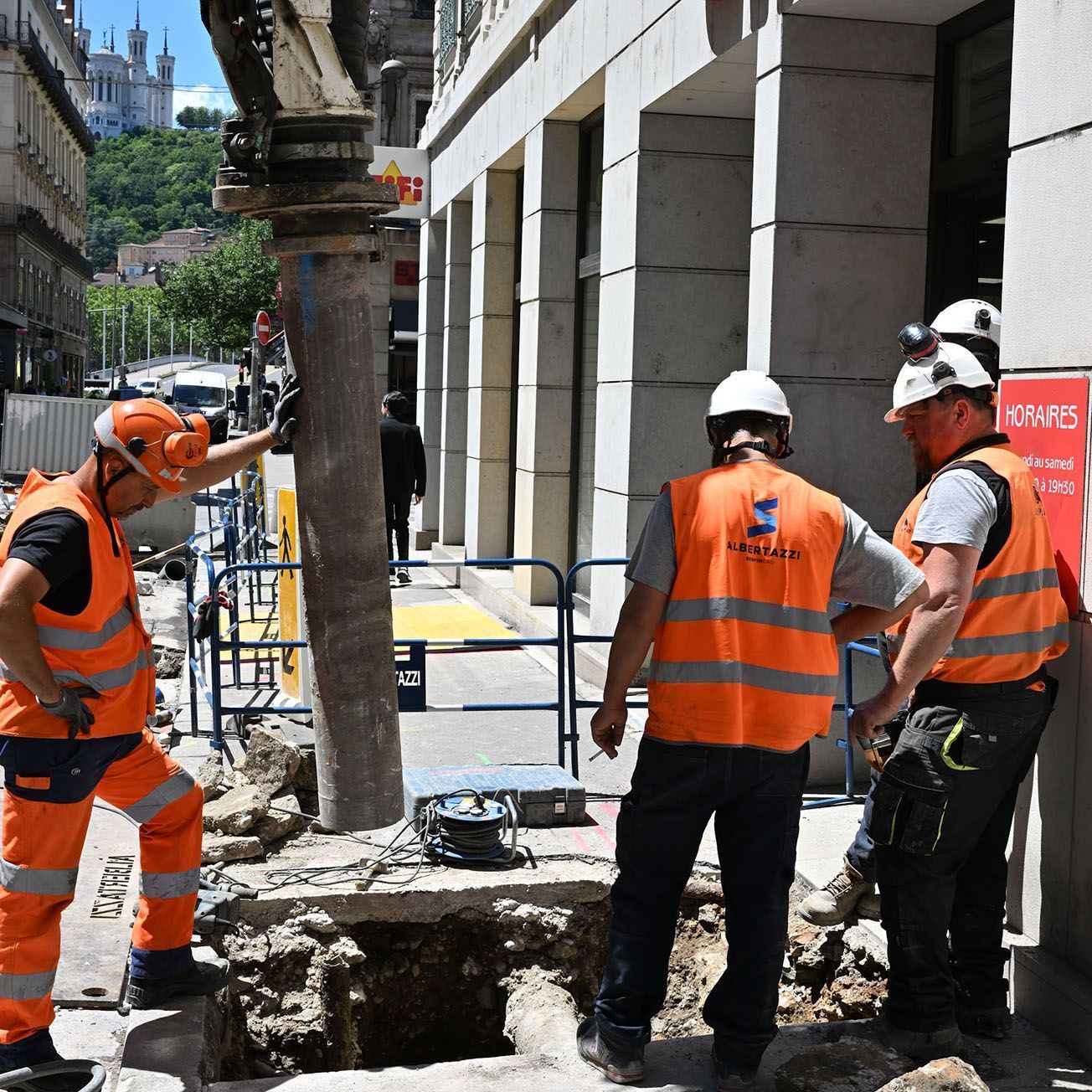 Trois ouvriers du bâtiment vêtus de gilets orange inspectent une tranchée de trottoir à côté d'un bâtiment.