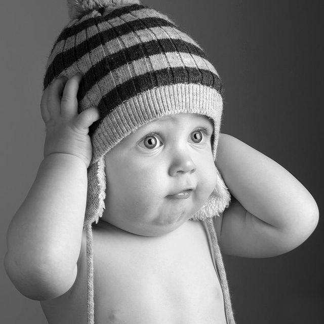 Bébé tenant un bonnet d'hiver rayé sur ses oreilles, les yeux grands ouverts sur fond uni.
