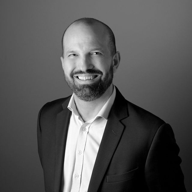 Homme souriant en costume sombre et chemise blanche sur fond gris uni.