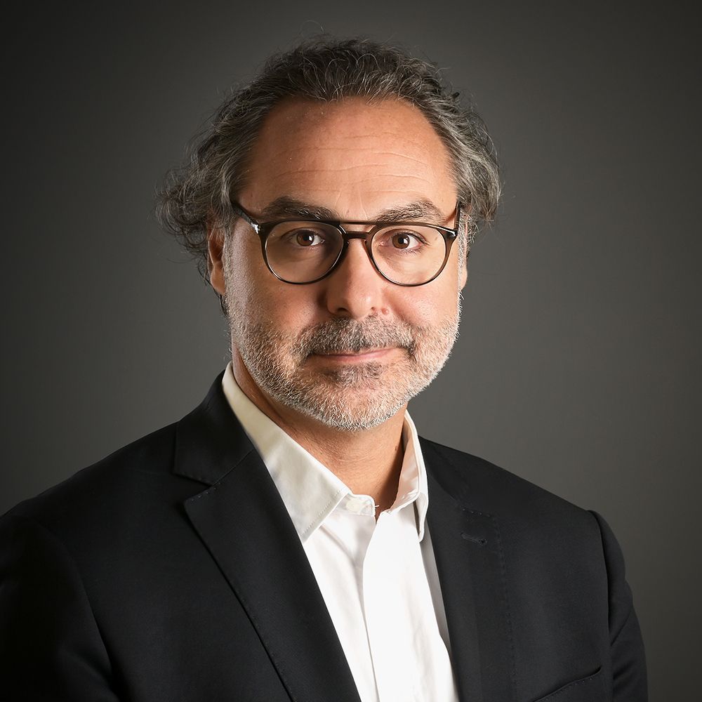 Portrait d'un homme en costume sombre et lunettes sur fond gris.
