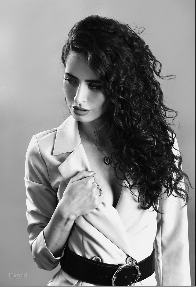 Portrait en noir et blanc d'une femme aux cheveux bouclés, vêtue d'un blazer et d'une ceinture, le regard pensif baissé.