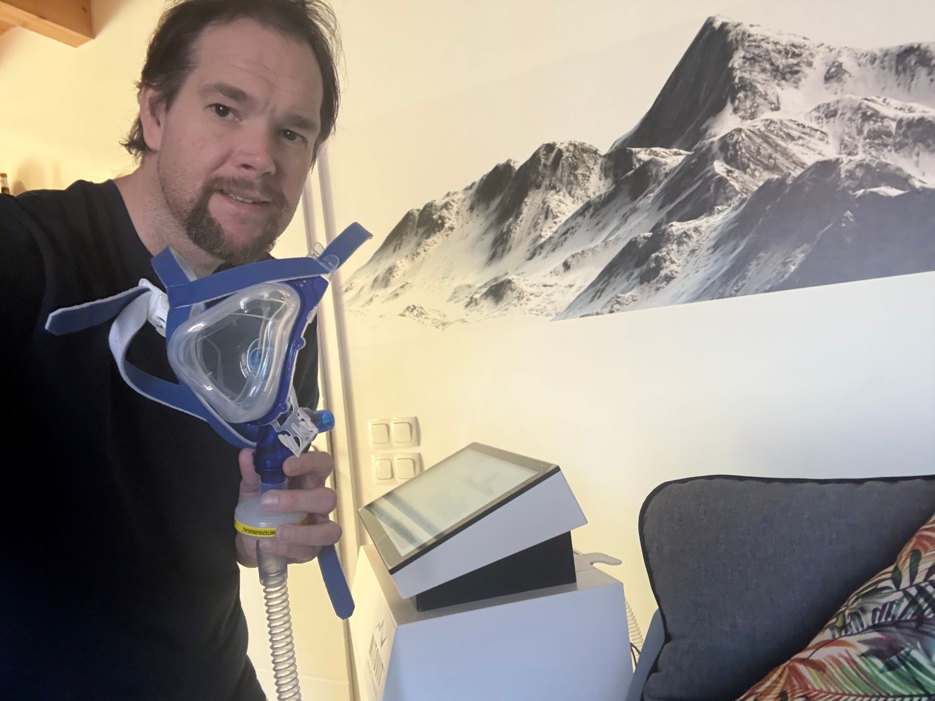 Ein Mann hält eine CPAP-Maske vor einem Bergbild an der Wand.