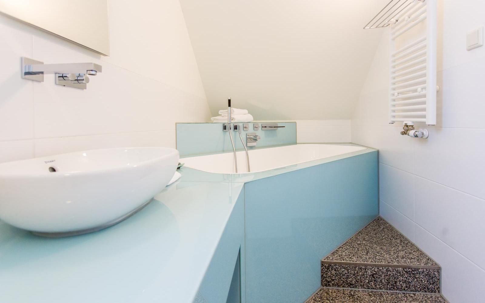 Ein modernes Badezimmer mit einem weißen Aufsatzwaschbecken auf hellblauer Arbeitsplatte, einer eingebauten Badewanne und Steinstufen.