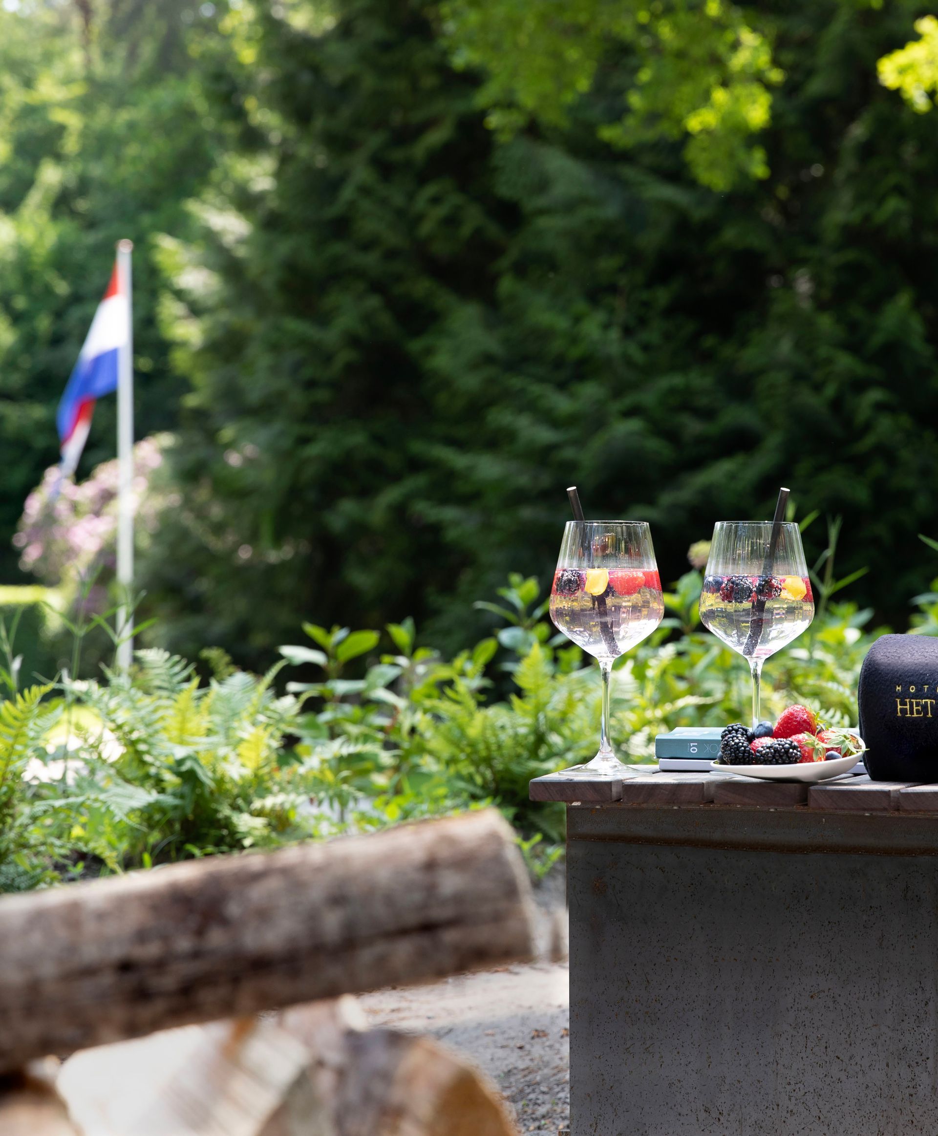 Een eettafel gedekt voor een maaltijd in de buitenlucht onder twee witte parasols op een gazon voor een huis met donkerhouten luiken.