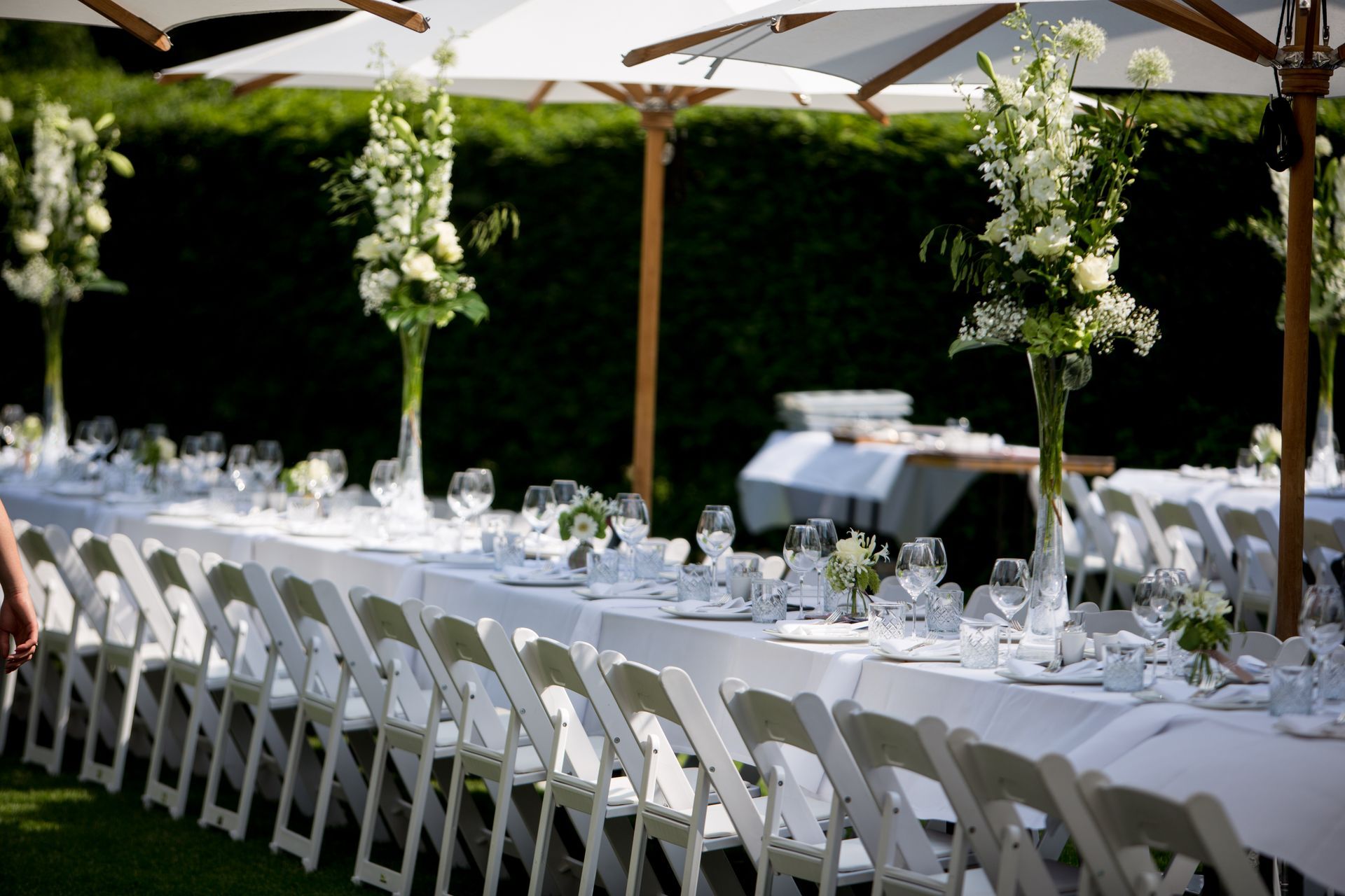 Lange buitentafels met witte tafelkleden en stoelen, gedekt voor een evenement en versierd met hoge witte bloemstukken.